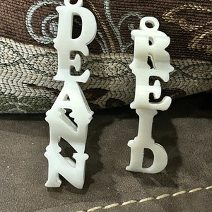 May include: Two white, decorative name pendants. One pendant spells out the name "DEAN" in a stylized font, and the other spells out "REED". Both pendants have a small loop at the top for hanging.