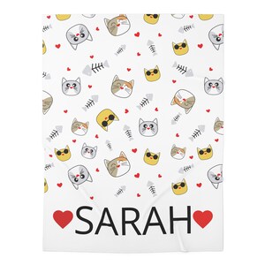 Op de afbeelding: Witte deken met een herhalend patroon van cartoon kattengezichten, visgraten en rode harten. De naam "SARAH" is in het zwart gedrukt met een rood hart aan weerszijden. De katten zijn in verschillende kleuren en stijlen.