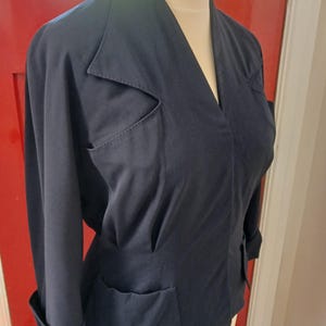 May include: A dark navy blue jacket featuring a V-neckline, pointed lapels, and long sleeves with folded cuffs. The garment includes two distinct chest pockets with angular flaps and two lower pockets, all constructed from a solid-coloured fabric.