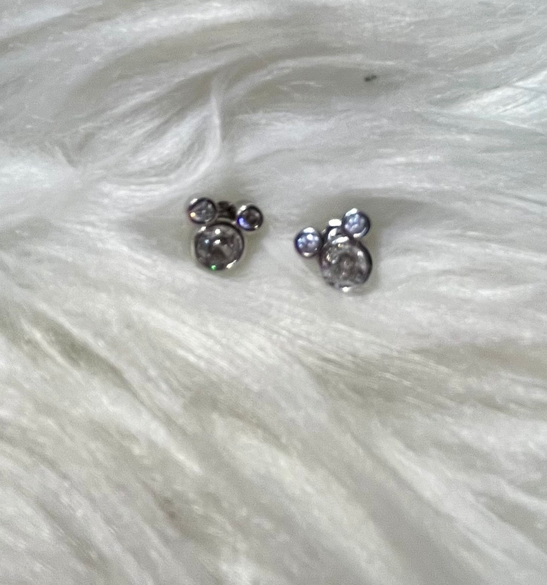 May include: A pair of silver earrings with clear crystal stones in the shape of Mickey Mouse heads.