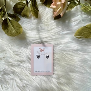 May include: A pair of silver stud earrings featuring a black and white Mickey Mouse design. The earrings are presented on a white card with the text "Our Boutique Girl" and a butterfly graphic.