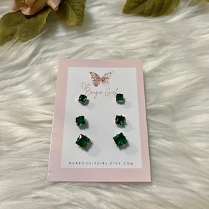 May include: A set of three pairs of square-cut, emerald-green stud earrings on a pink card. The card has the text "Our Bougie Girl" and the Etsy website address. The earrings are displayed on a white, fluffy surface.