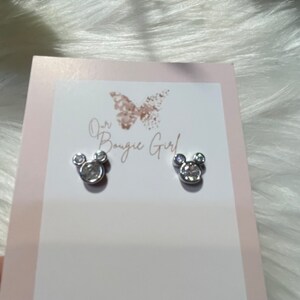 May include: A pair of silver earrings shaped like Mickey Mouse heads with clear crystal accents. The earrings are displayed on a white card with the text "Our Bougie Girl".