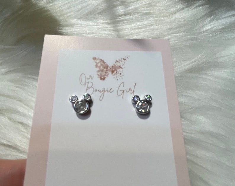 May include: A pair of silver earrings shaped like Mickey Mouse heads with clear crystal accents. The earrings are displayed on a white card with the text "Our Bougie Girl".