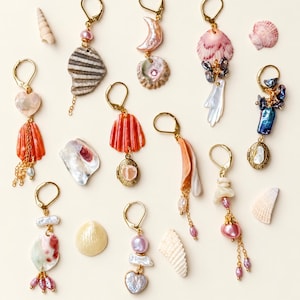 May include: A collection of gold-tone leverback earrings featuring various seashells, pearls, and small metal lockets. The designs incorporate pink, white, blue, and orange hues with dangling chains and charms, arranged alongside loose individual seashells.