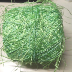 May include: A square-shaped ball of light green yarn featuring a shiny, metallic finish. The yarn consists of multiple thin, interwoven strands with loose, wispy fibers extending from the main body of the ball, creating a textured appearance.