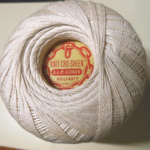 May include: A ball of off-white cotton crochet thread wound in a spherical shape. The center features a circular yellow label with red text reading "KNIT-CRO-SHEEN", "J. & P. COATS", "BOILFAST", and "U.S.A." surrounded by a red chain link graphic.