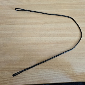 Puede incluir: Un cordón negro trenzado con lazos en ambos extremos, sobre una superficie de madera marrón claro. El cordón es fino y parece estar hecho de un material tejido. Los lazos son pequeños y simétricos.