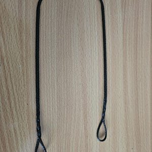 Puede incluir: Un cordón negro trenzado con un lazo en cada extremo. El cordón tiene forma de U sobre una superficie de madera marrón claro. Los lazos son pequeños y parecen estar anudados o fusionados.