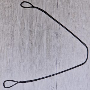 Puede incluir: Un cordón negro con lazos en ambos extremos, sobre una superficie clara con vetas de madera. El cordón parece estar hecho de un material tejido, y los lazos se forman anudando el cordón.