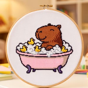May include: Cross-stitch embroidery in a wooden hoop features a brown capybara in a pink bathtub with yellow rubber duckies. The capybara is surrounded by white bubbles. The text "MOCKUP IMAGE FOR DISPLAY PURPOSES ONLY" is at the bottom.