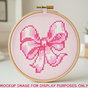 May include: A cross-stitch embroidery hoop featuring a pink bow design. The bow is rendered in shades of pink and white against a light pink background. The wooden hoop is secured with a brass screw. The text "MOCKUP IMAGE FOR DISPLAY PURPOSES ONLY" is at the bottom.