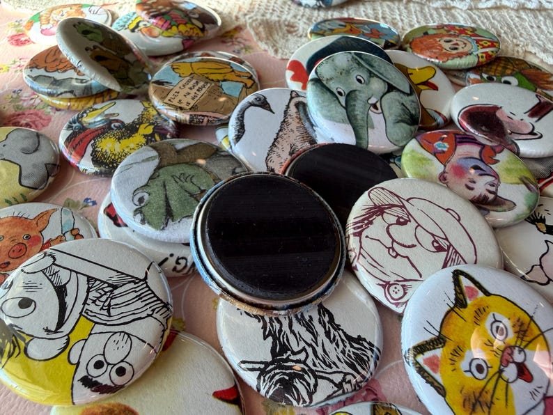 May include: A collection of round magnets with various cartoon illustrations. The magnets feature images of animals, people, and other whimsical designs in bright colors. Some magnets have a glossy finish and a black magnetic backing.