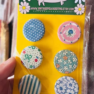 May include: Set of six round pinback buttons with various patterns, including polka dots, floral designs, and stripes. The buttons are displayed on a yellow backing card with a pig illustration and the website address www.vintagepolkadotpig.etsy.com.