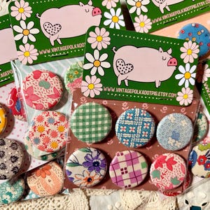 May include: A collection of colorful fabric-covered buttons with various patterns, including floral, gingham, and geometric designs. The buttons are displayed with green cards featuring a pig illustration and the website address vintagepolkadotpig.etsy.com.