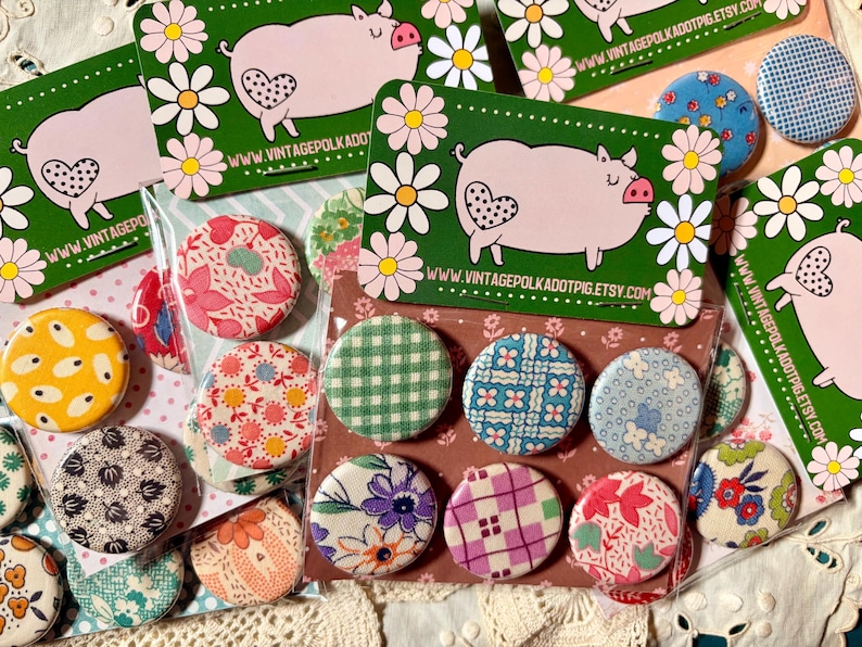 May include: A collection of colorful fabric-covered buttons with various patterns, including floral, gingham, and geometric designs. The buttons are displayed with green cards featuring a pig illustration and the website address vintagepolkadotpig.etsy.com.