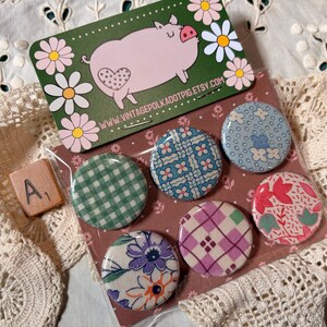 May include: A set of six round fabric-covered buttons with various floral and geometric patterns. A green card with a pink pig illustration and a wooden tile with the letter "A" and the number "1" are also visible.