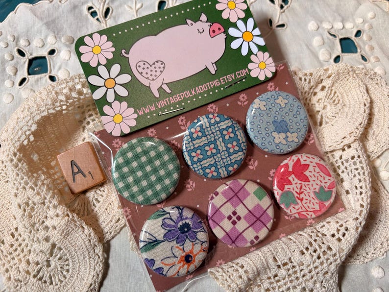 May include: A set of six round fabric-covered buttons with various floral and geometric patterns. A green card with a pink pig illustration and a wooden tile with the letter "A" and the number "1" are also visible.