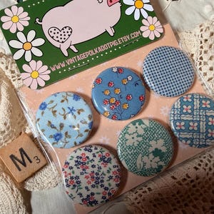 May include: A set of six round fabric-covered buttons with floral and patterned designs in blue, white, and red. The buttons are packaged with a pig-themed card and a Scrabble tile.
