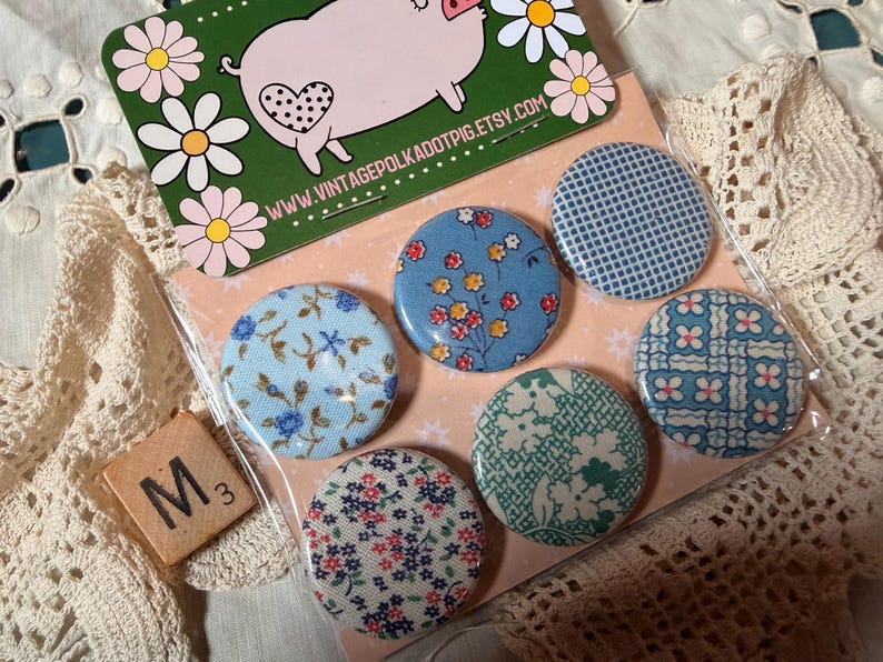 May include: A set of six round fabric-covered buttons with floral and patterned designs in blue, white, and red. The buttons are packaged with a pig-themed card and a Scrabble tile.