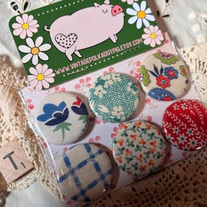 May include: A set of six fabric-covered buttons with various floral and geometric patterns. The buttons are in a clear plastic package, with a green card featuring a pig illustration and the text "www.vintagepolkadotpig.etsy.com".