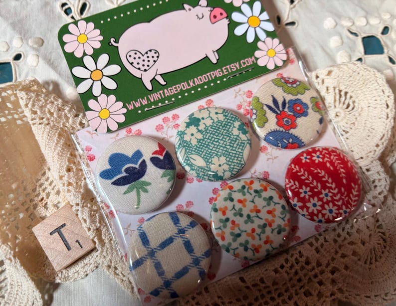 May include: A set of six fabric-covered buttons with various floral and geometric patterns. The buttons are in a clear plastic package, with a green card featuring a pig illustration and the text "www.vintagepolkadotpig.etsy.com".
