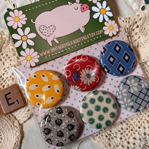 May include: A set of six round, patterned buttons in a clear plastic package. The buttons feature various designs, including floral, geometric, and abstract patterns in colors like red, blue, yellow, and green. A pig-themed card and a tile with the letter "E" are also visible.