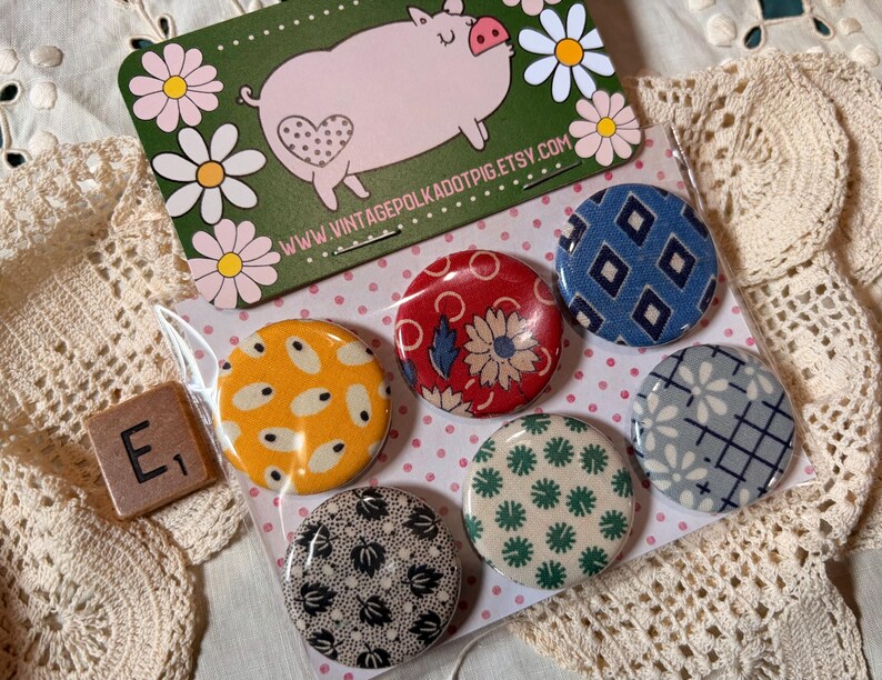 May include: A set of six round, patterned buttons in a clear plastic package. The buttons feature various designs, including floral, geometric, and abstract patterns in colors like red, blue, yellow, and green. A pig-themed card and a tile with the letter "E" are also visible.