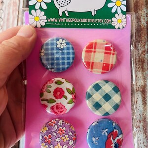 May include: A set of six round fabric-covered buttons with various floral and plaid patterns. The buttons are packaged on a pink backing card, with a green header featuring a pig illustration and the website address www.vintagepolkadotpig.etsy.com.