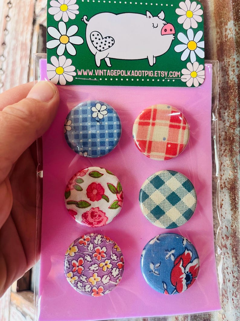 May include: A set of six round fabric-covered buttons with various floral and plaid patterns. The buttons are packaged on a pink backing card, with a green header featuring a pig illustration and the website address www.vintagepolkadotpig.etsy.com.