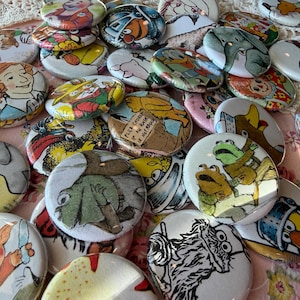 May include: A collection of colorful, round buttons with various cartoon illustrations. The buttons feature images of animals, characters, and text. The buttons are scattered on a pink floral surface.