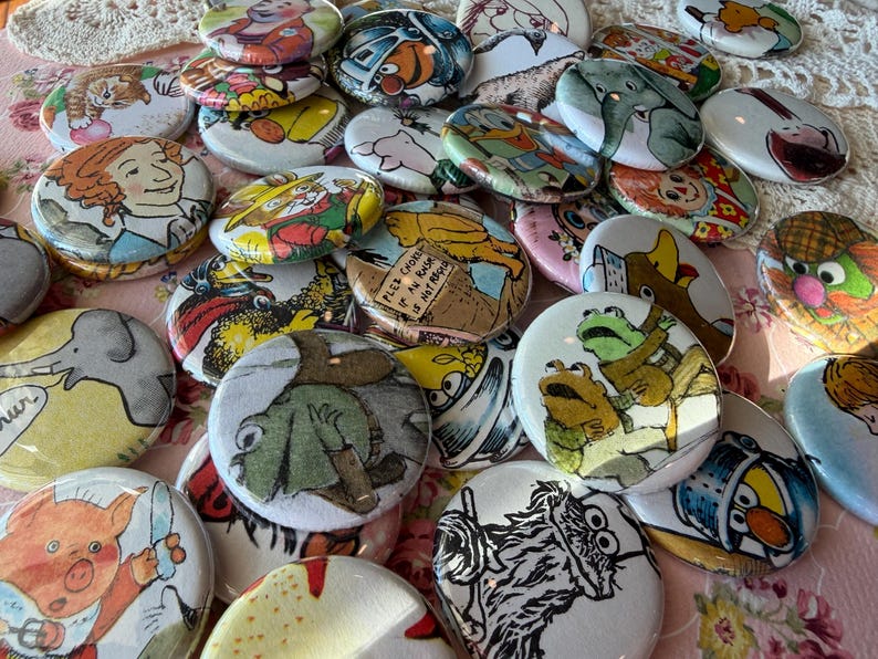 May include: A collection of colorful, round buttons with various cartoon illustrations. The buttons feature images of animals, characters, and text. The buttons are scattered on a pink floral surface.