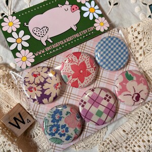 May include: A collection of six round fabric-covered buttons with various floral and plaid patterns. A green business card with a pig illustration and the text "www.vintagepolkadotpig.etsy.com" is visible. A wooden tile with the letter "W" is also present.