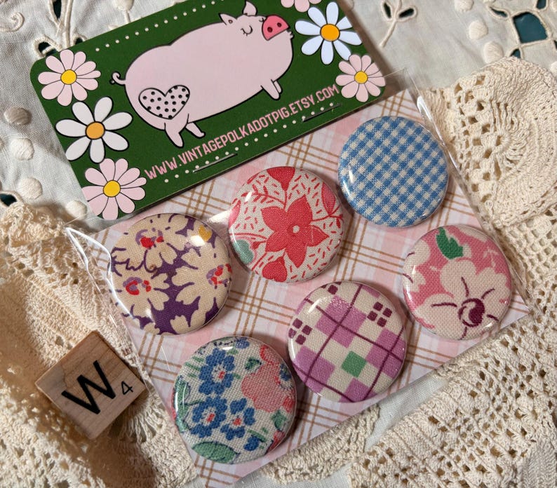 May include: A collection of six round fabric-covered buttons with various floral and plaid patterns. A green business card with a pig illustration and the text "www.vintagepolkadotpig.etsy.com" is visible. A wooden tile with the letter "W" is also present.