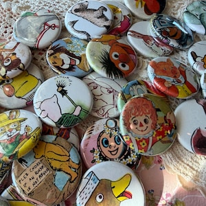 May include: A collection of colorful, round pinback buttons featuring various cartoon characters and illustrations. The buttons showcase images of animals, people, and other whimsical designs. The buttons are arranged on a textured, off-white surface.