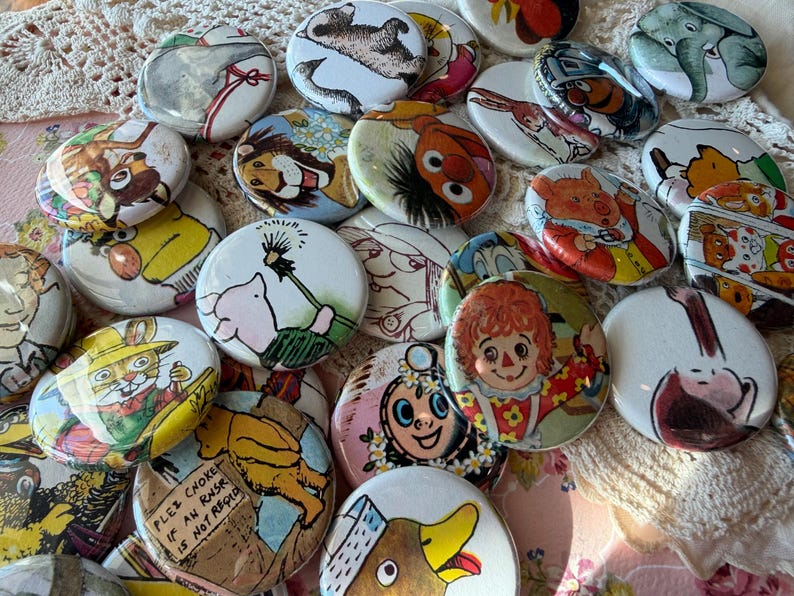 May include: A collection of colorful, round pinback buttons featuring various cartoon characters and illustrations. The buttons showcase images of animals, people, and other whimsical designs. The buttons are arranged on a textured, off-white surface.