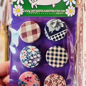 May include: A set of six round fabric-covered buttons with various patterns, including gingham, floral, and polka dots. The buttons are displayed on a purple background, with a pig illustration at the top. The website address www.vintagepolkadotpig.etsy.com is visible.