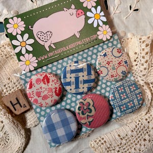 May include: A collection of six vintage-style pinback buttons with various patterns, including floral, gingham, and geometric designs. A business card with a pig illustration and the text "Vintage Polka Dot Pig" is also present. The buttons are on a polka dot background.