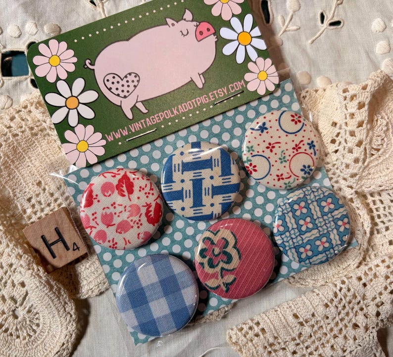 May include: A collection of six vintage-style pinback buttons with various patterns, including floral, gingham, and geometric designs. A business card with a pig illustration and the text "Vintage Polka Dot Pig" is also present. The buttons are on a polka dot background.