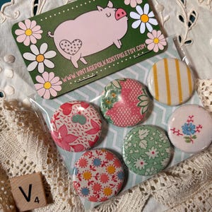 May include: A collection of six round pinback buttons with various floral and patterned designs. A green card with a pig illustration and the text "www.vintagepolkadotpig.etsy.com" is also visible. A wooden tile with the letter "V" and the number "4" is in the image.