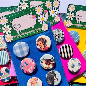 May include: A collection of colorful pinback buttons with various designs, including floral patterns, gingham, and vintage illustrations. The buttons are arranged on colorful cardstock and packaged with a pig graphic.