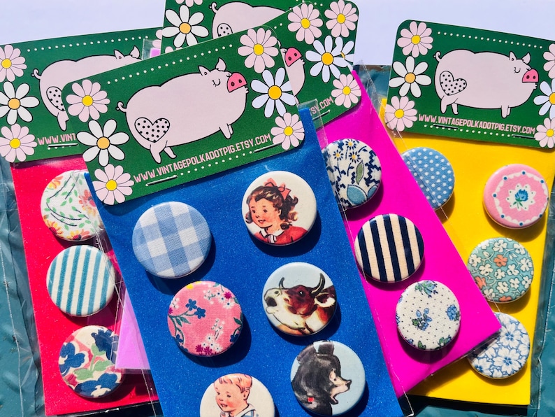 May include: A collection of colorful pinback buttons with various designs, including floral patterns, gingham, and vintage illustrations. The buttons are arranged on colorful cardstock and packaged with a pig graphic.