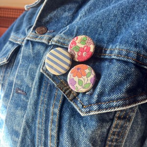 May include: Three round fabric buttons pinned to a denim jacket. One button features blue and white stripes, while the others display floral patterns in pink, orange, and purple. The jacket is a medium wash blue.