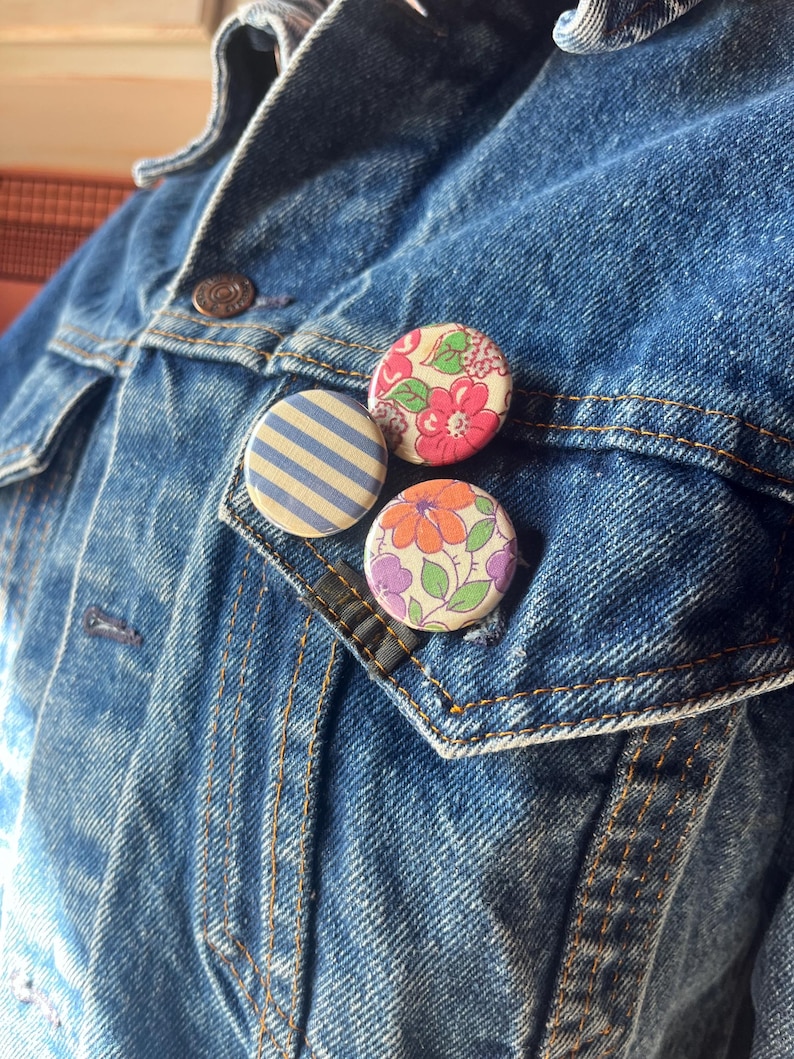 May include: Three round fabric buttons pinned to a denim jacket. One button features blue and white stripes, while the others display floral patterns in pink, orange, and purple. The jacket is a medium wash blue.