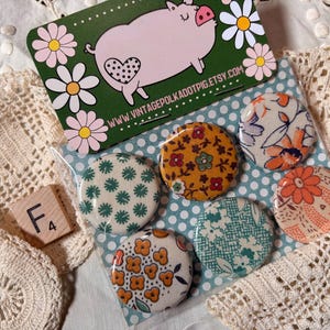 May include: A collection of six round, patterned buttons in a clear plastic sleeve. The buttons feature floral and geometric designs in various colors. A business card with a pig illustration and the text "www.vintagepolkadotpig.etsy.com" is also visible.