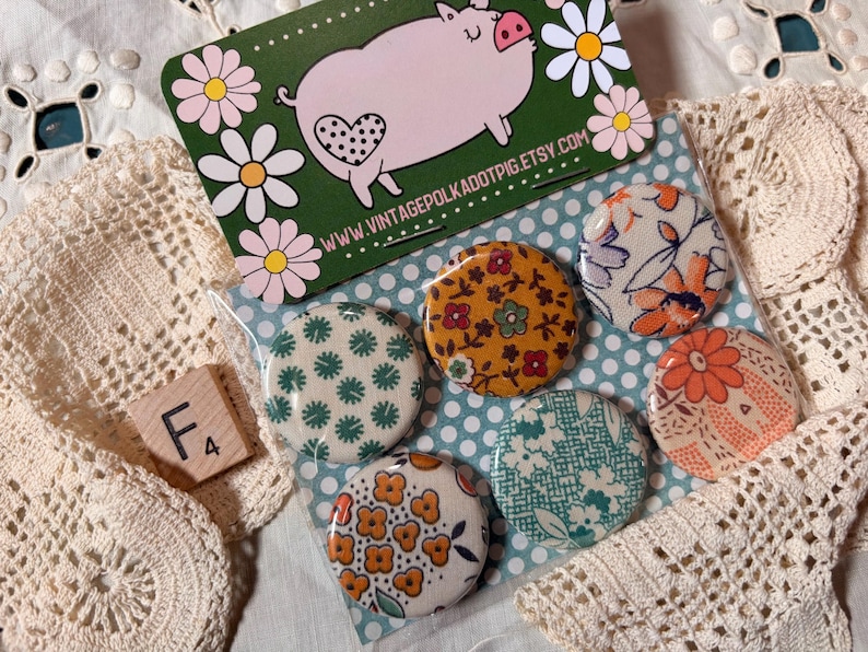 May include: A collection of six round, patterned buttons in a clear plastic sleeve. The buttons feature floral and geometric designs in various colors. A business card with a pig illustration and the text "www.vintagepolkadotpig.etsy.com" is also visible.