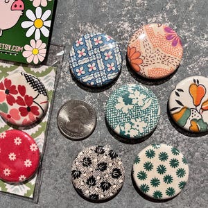 May include: A collection of fabric-covered buttons with various floral and geometric patterns in red, blue, green, and orange. A quarter coin is next to the buttons. The buttons are on a gray surface.