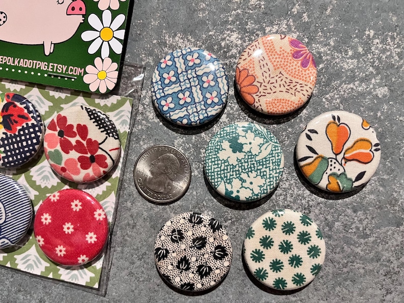 May include: A collection of fabric-covered buttons with various floral and geometric patterns in red, blue, green, and orange. A quarter coin is next to the buttons. The buttons are on a gray surface.