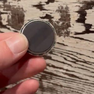 May include: A round magnet with a dark gray surface and a silver-colored rim. The magnet is held in a person's hand against a weathered, light-colored wooden surface.