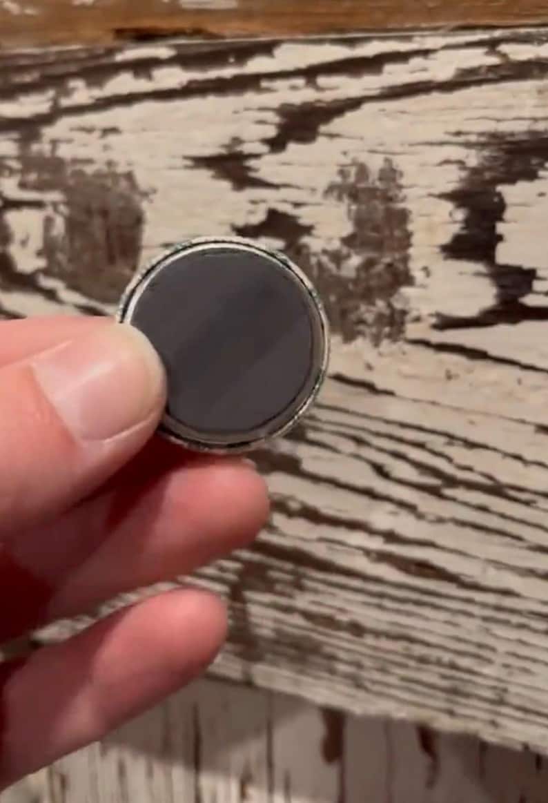 May include: A round magnet with a dark gray surface and a silver-colored rim. The magnet is held in a person's hand against a weathered, light-colored wooden surface.